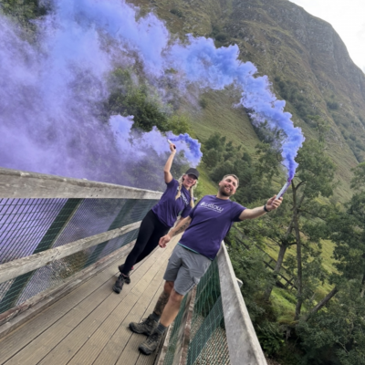 Colleague and partner holding purple flares at top of mountain during recent challenge