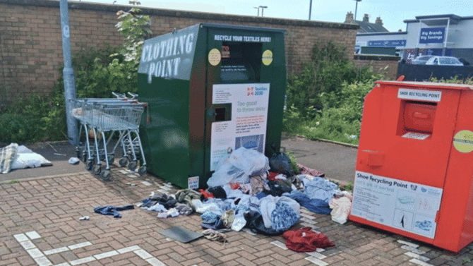 Two charity clothing banks, with shops in the background and burst bags of donations strewn in the foreground