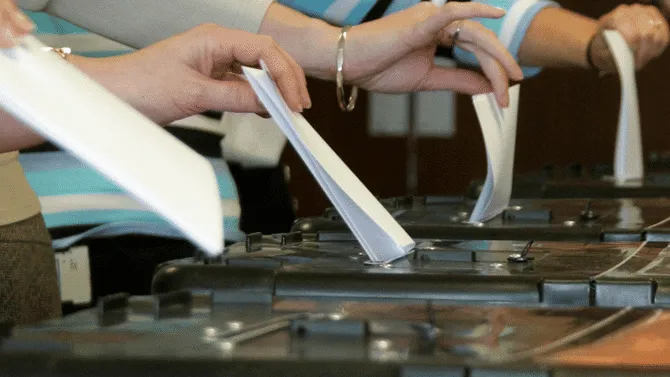 Four people's hands placing ballot papers into a ballot box during an election