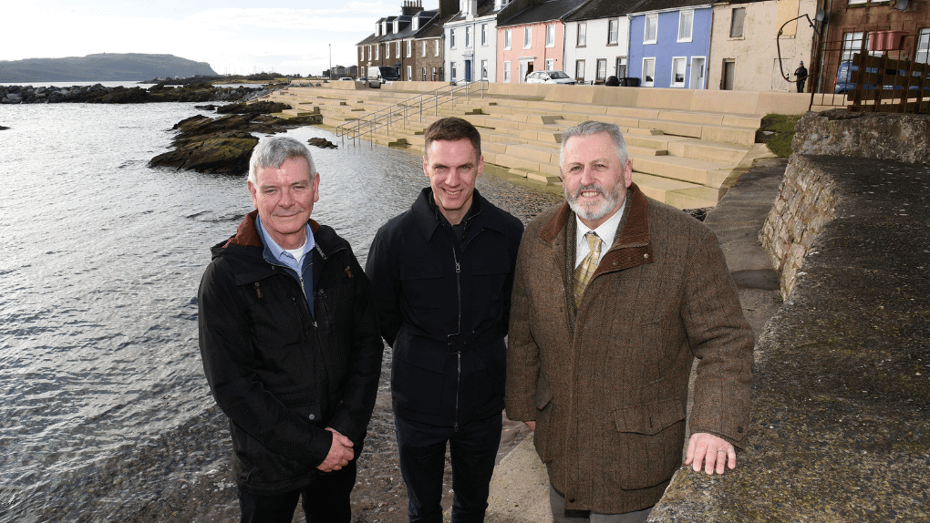 Three men standing in a row near to the shore with colourful houses in the background