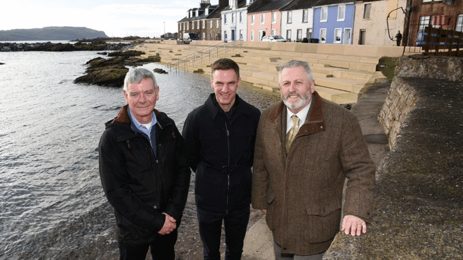 Three men standing in a row near to the shore with colourful houses in the background
