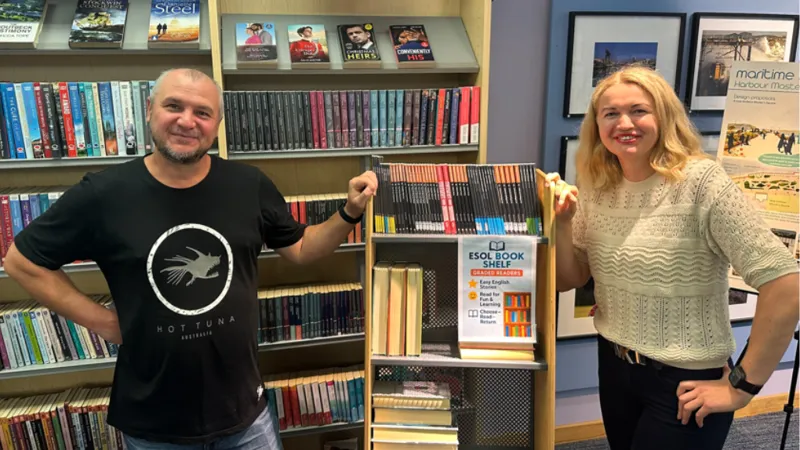 A man and woman standing beside the English for Speakers of Other Languages book shelf in Irvine library